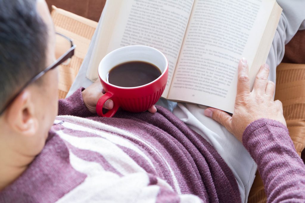 Reclining while reading a book with hot coffee in red coffee cup in right hand in a warm and relaxing home atmosphere.
